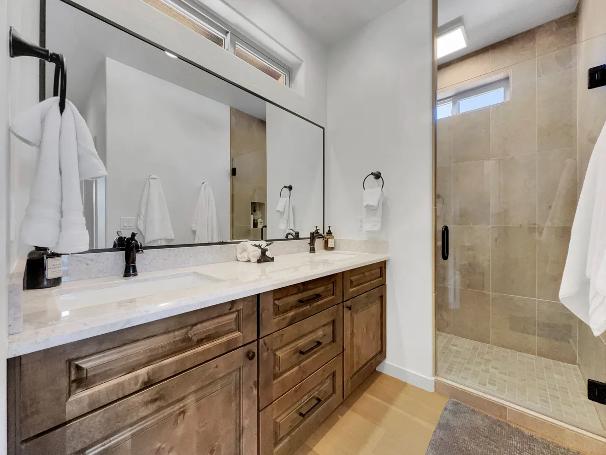 A spacious, modern bathroom featuring a double sink vanity with a large mirror, walk-in shower, wooden cabinetry, and fresh white towels. The space is organized, serene, and spa-like, inviting guests to unwind with refreshing morning or evening routines.