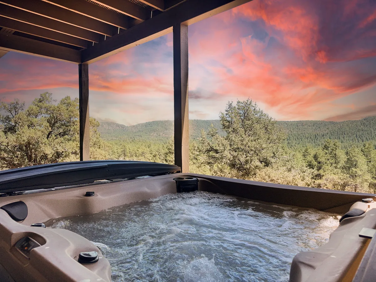 This image captures a private hot tub set on a wooden deck, overlooking scenic mountain views beneath a striking sunset sky. The tranquil and immersive forest setting is enhanced by the warm colors of dusk, and the hot tub is positioned for ultimate privacy and relaxation.