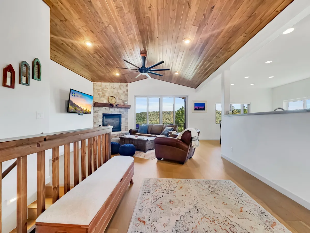 A spacious, welcoming great room featuring a soaring wooden vaulted ceiling, a striking stone fireplace, comfortable seating, and expansive windows framing scenic forest views. Elements of rustic charm blend seamlessly with modern comfort, and the room is bathed in natural light.