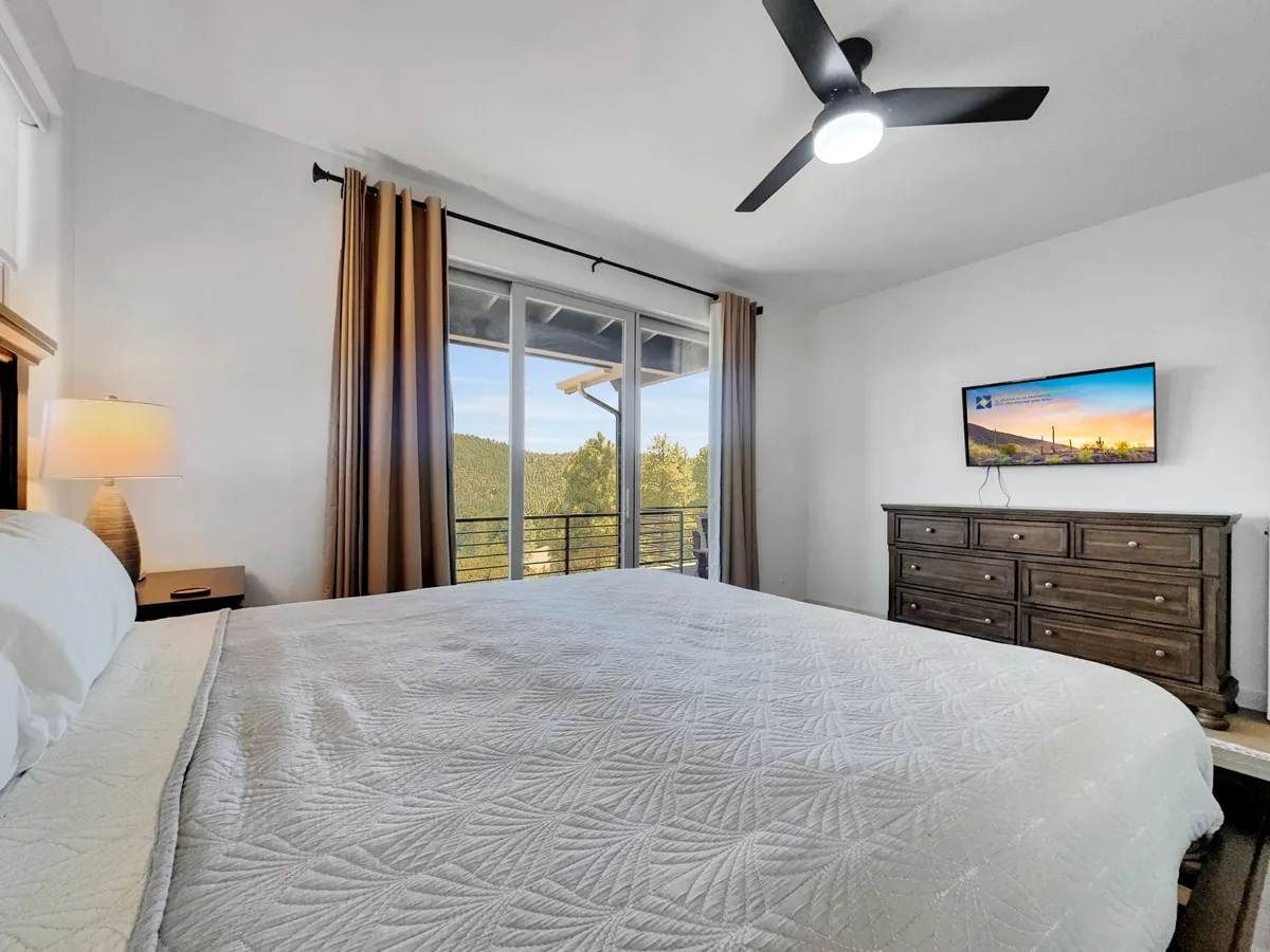 This serene bedroom features a bed with quilted bedding, large windows showcasing a mountain view, a wooden dresser, and a TV mounted on the wall. The room is filled with natural light, creating an inviting atmosphere perfect for relaxation or enjoying the view from bed.