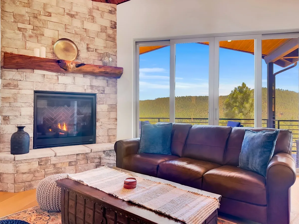 A warm and welcoming living area featuring a cozy fireplace, a comfortable sofa, and expansive windows framing scenic outdoor views. The space invites groups to unwind by the fire, share laughter, and watch sunsets together.