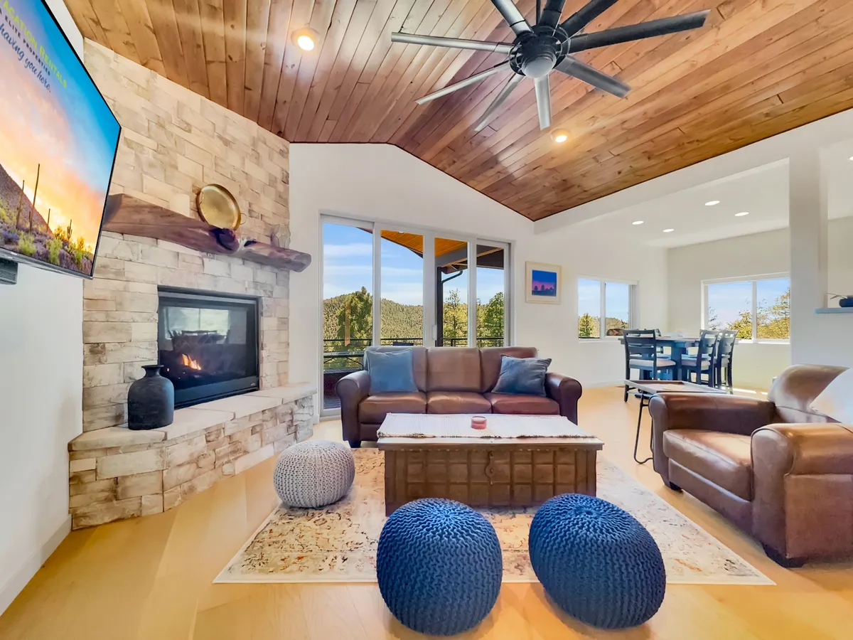 A cozy living area with a stone fireplace, leather sofa, wooden ceiling, decorative poufs, and a view into the adjacent dining space. Sunlight streams through the windows, highlighting the inviting, multi-use character of the great room.