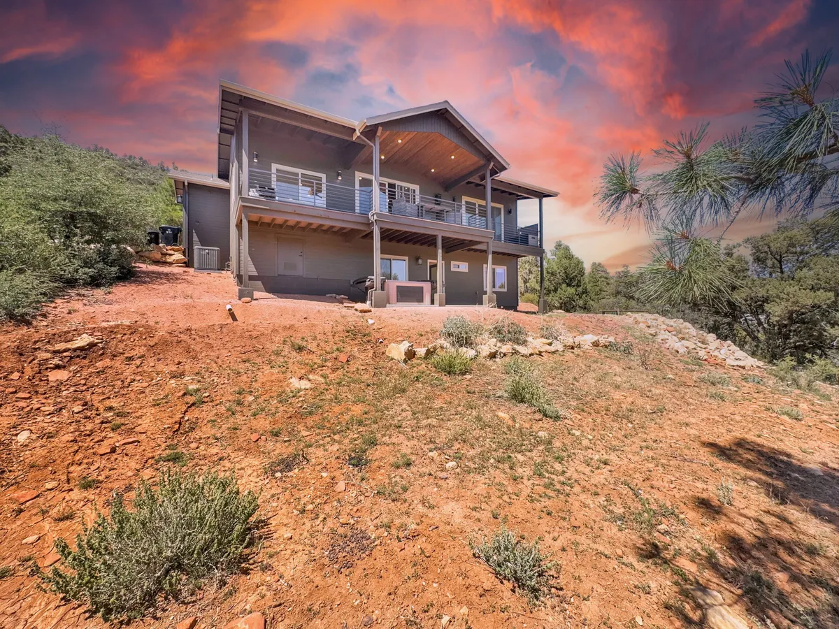 A charming exterior view of the house nestled in Arizona's high country, surrounded by scenic landscape and an expansive sky. The porch and balcony invite outdoor gatherings, while the property hints at proximity to hiking trails and nature activities.