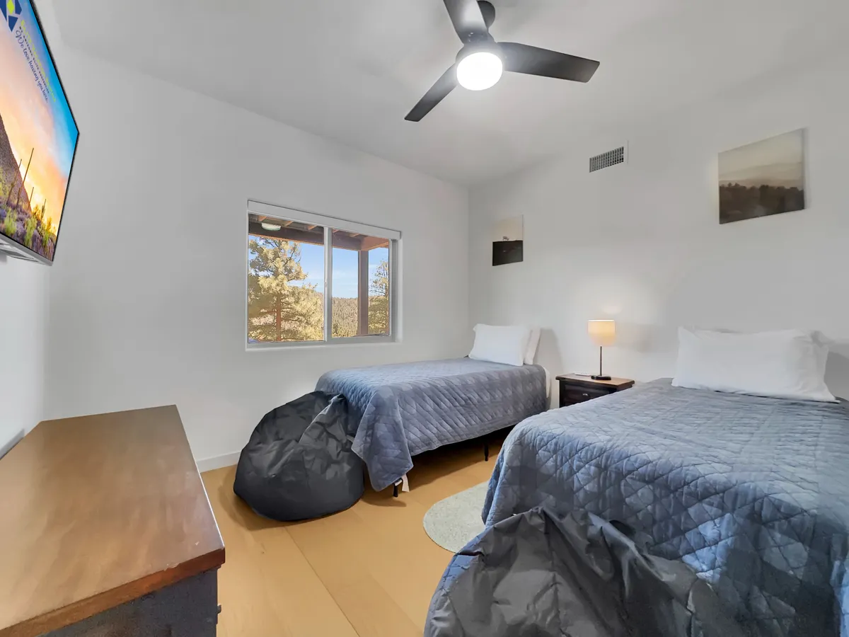 A cozy bedroom equipped with twin beds, a TV mounted on the wall, a window with a relaxing view, and a nightstand with a lamp. The space is comfortable and bright, inviting guests to relax, watch TV, or enjoy the outdoors through the window.